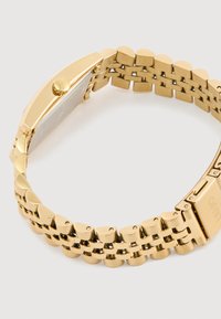 Χρυσής απόχρωσης ρολόι χειρός με συνδεδεμένο λουράκι, επίπεδη επιφάνεια και υφασμάτινη ανάγλυφη διακόσμηση. Διαθέτει στρογγυλή θήκη και λεία, γυαλιστερή φινίρισμα.