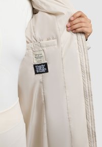 Cappotto beige isolante con finitura satinata, dotato di interno trapuntato, etichette per la cura e un tag del marchio visibile all'interno della tasca.