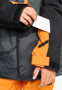 Veste noire et orange avec une poche zippée, motif gris texturé, poignets en velcro, et une main insérant une carte dans la poche de la manche.
