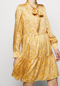 Robe jaune à motifs avec un col à nœud papillon, des manches longues et une jupe à volants. Fabriquée en tissu léger avec une texture plissée.