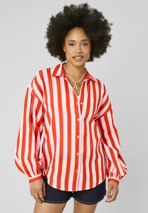 Jeune femme avec coiffure afro portant un t-shirt à manches longues rayé vertical rouge et blanc, des shorts foncés et un collier en forme d'étoile de mer, debout devant un fond gris.