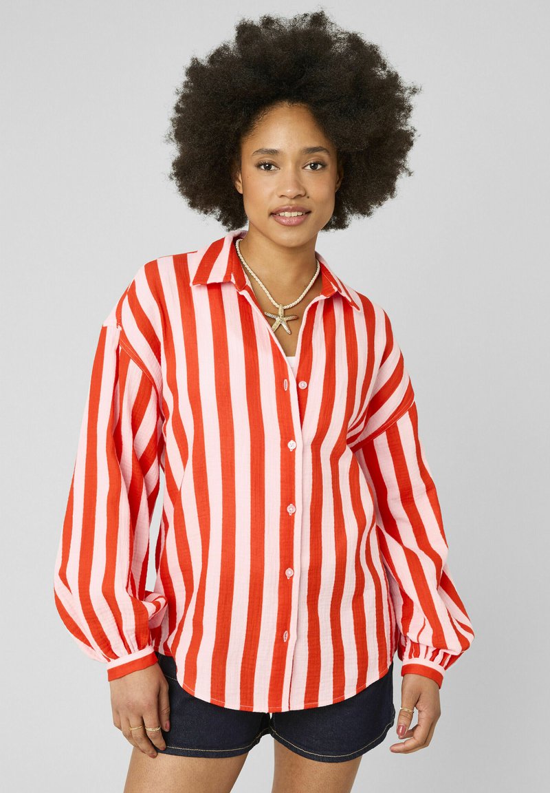 Jeune femme avec coiffure afro portant un t-shirt à manches longues rayé vertical rouge et blanc, des shorts foncés et un collier en forme d'étoile de mer, debout devant un fond gris.
