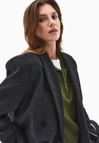 Musta villablazer, jossa on teksturoidu pinta, ylipitkä istuvuus ja suuret napit, päällystetty vihreän vetoketjullisen neuleen päällä.