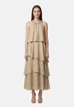 Donna in piedi che indossa un abito lungo beige senza maniche a balze con scollatura halter decorata e sandali beige con tacchi alti.