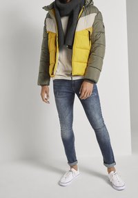 Giacca imbottita con colori verde oliva, grigio chiaro e giallo. Presenta un cappuccio, chiusura con zip e una sciarpa grigia strutturata. Indossata con jeans blu e sneakers bianche.