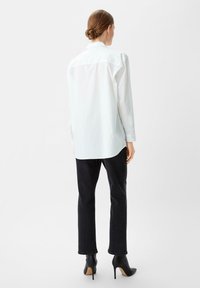 Witte button-up shirt met lange mouwen, ontspannen pasvorm en een licht gebogen zoom. Gecombineerd met zwarte rechte jeans en zwarte hoge hakken.