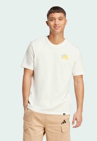 White cotton t-shirt with a round neckline, short sleeves, and yellow graphic detail on the left chest. Worn with beige cargo pants.