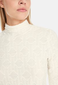 White, textured high-neck top featuring a geometric pattern with raised lines and diamond shapes. Fitted design, smooth fabric finish.