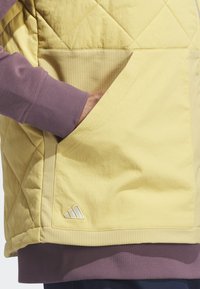 Žltá prešívaná vesta s rebrovanými fialovými manžetami a postrannými panelmi. Obsahuje horizontálne pruhovanú kapsu a malú akcent kov na logu Adidas.