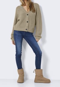 Beige stickad kofta med V-ringning, fyra knappar och oversize ärmar, kombinerad med blå skinny jeans och ljusbruna pufferskor.