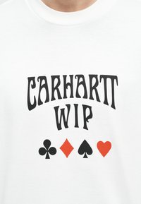 Λευκό πουκάμισο με μαύρο κείμενο «CARHARTT WIP» πάνω από τέσσερα σύμβολα τραπουλόχαρτων: μαύρο κλαρί, κόκκινο ρόμβο, μαύρο σπαθί και κόκκινη καρδιά.