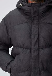 Doudoune noire avec un motif chevron texturé, munie d'une fermeture éclair frontale, d'une capuche ajustable et de poches latérales.