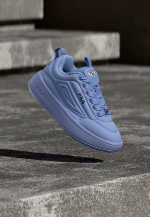 Light blue Fila sneaker with laces, floating above a concrete surface with stone steps in the background.