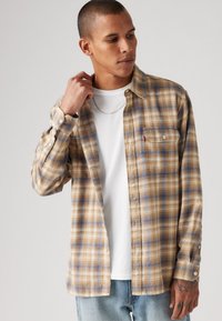 Camisa xadrez em tons de bege, azul e castanho; feita de um tecido macio com bolso no peito com botão e detalhes nas mangas ajustáveis. T-shirt branca por baixo.