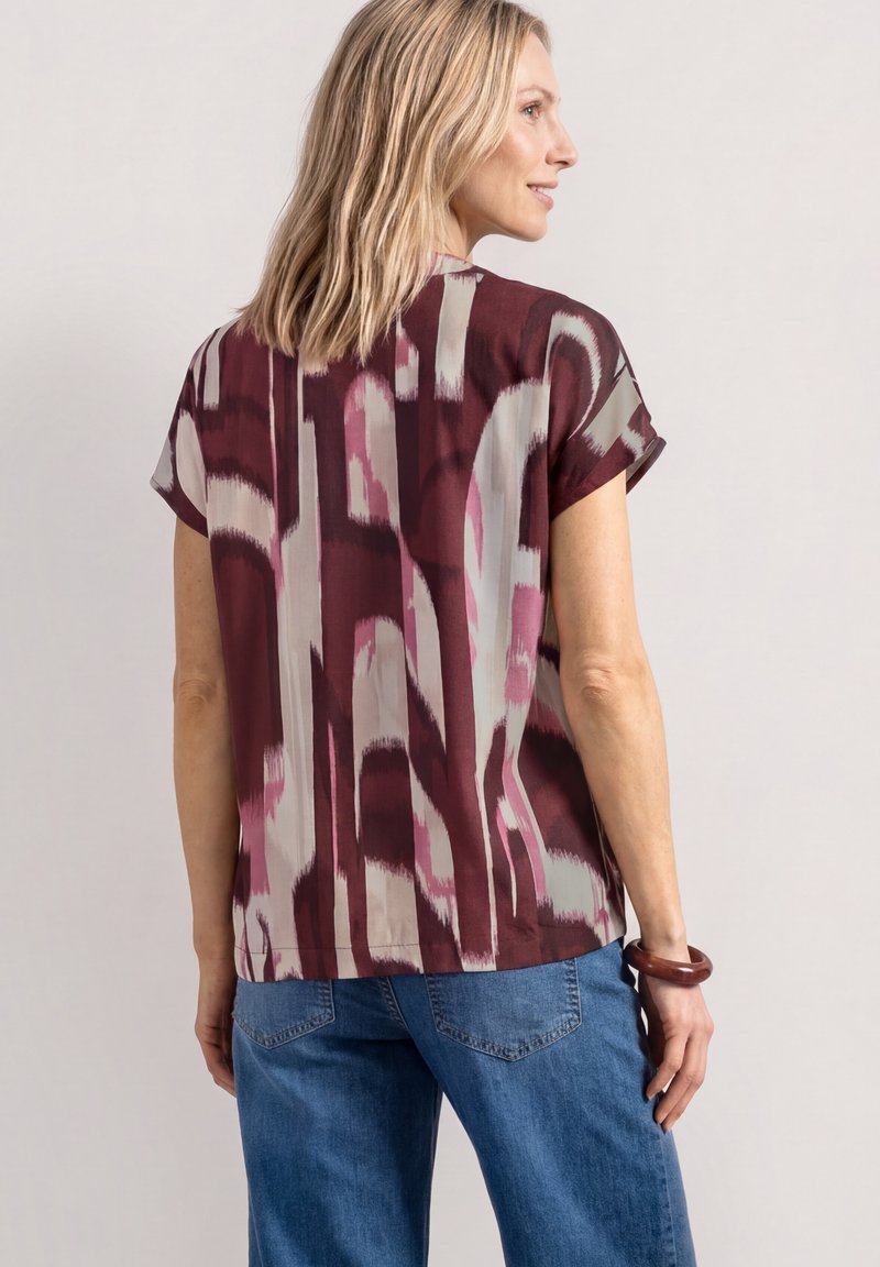 Femme aux cheveux blonds mi-longs portant un chemisier à manches courtes avec un motif abstrait bordeaux et beige et un jean bleu, debout de profil.