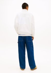 Λευκό μπουφάν από φλις με ψηλό γιακά και διακριτική λεπτομέρεια λογότυπου, συνδυασμένο με φαρδιά σκούρα μπλε παντελόνια από τζιν με τσέπες.