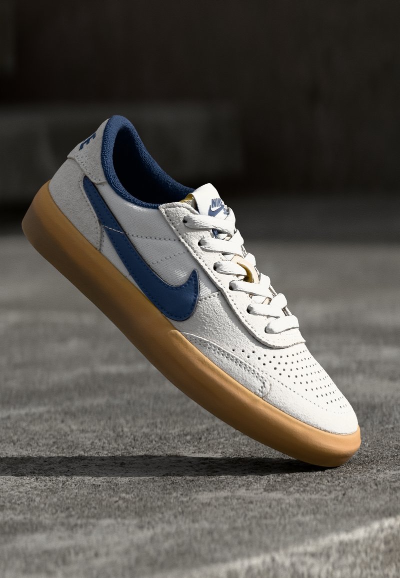 Valkoiset ja siniset Nike-tossut, joissa on kumipohja, leijuvat harmaalla teksturoidulla pinnalla pehmeässä valossa.