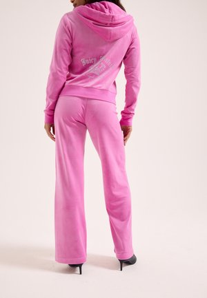 Donna che indossa una tuta Juicy Couture in velour rosa con logo in strass sulla schiena, abbinata a scarpe nere con tacco alto, vista di spalle.
