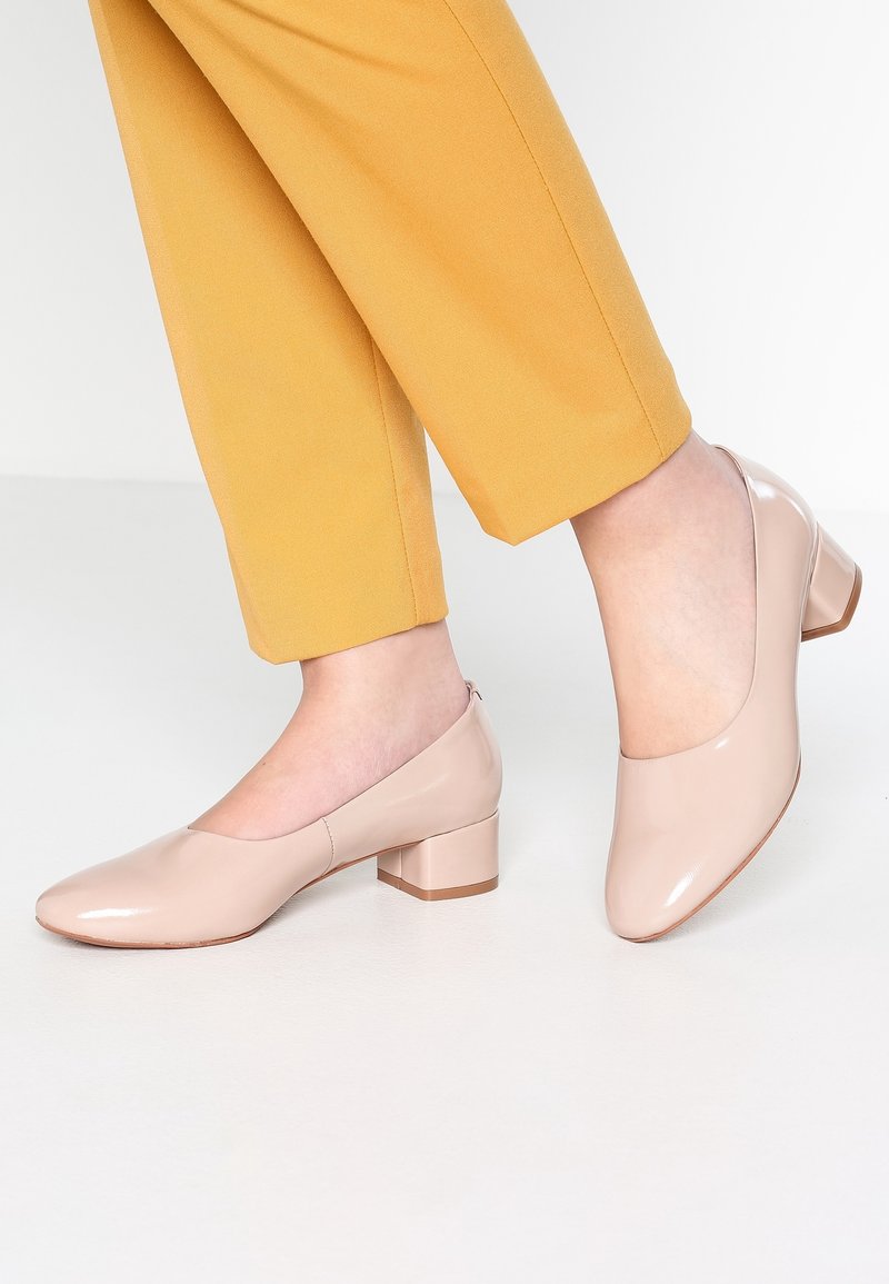 Pieds portant des chaussures brillantes beige à petit talon carré, associées à un pantalon jaune moutarde longueur cheville, sur un sol blanc.