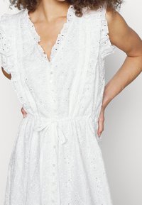 Vit klänning med volangärmar, knäppning framtill och dragsko i midjan. Har ett texturerat tyg med ögonhålsmönster och en avslappnad passform.