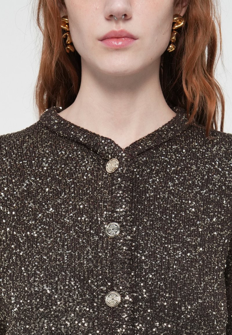 Frau mit roten Haaren, die eine dunkle, mit Pailletten besetzte Strickjacke mit drei verzierten silbernen Knöpfen und goldenen, gedrehten Creolen trägt.