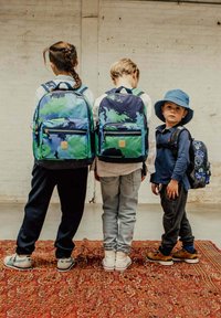 Drie kinderen met rugzakken: twee hebben grote rugzakken met blauw-groene camouflagepatronen, en één heeft een kleine zwarte rugzak met blauwe patronen.