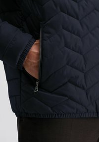 Marineblauwe gewatteerde jas met een ritszak aan de zijkant en elastische manchetten. De stof heeft een gladde textuur en een ruitvormig patroon.