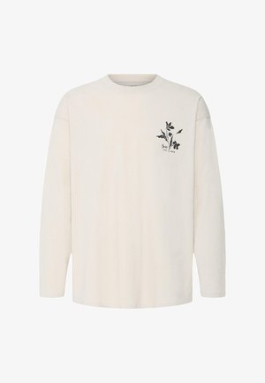 Garām piedurknēm bēšs krekls ar apaļu apkakli un nelielu melnu ziedu dizainu uz kreisā krūšu rajona, ieskaitot tekstu "Sanita EST. 1999".