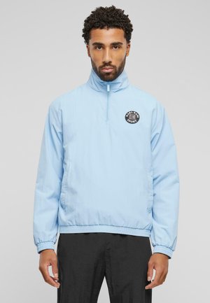 Lysblå halv-zip jakke med elastiske manchetter og kant, der har et sort rundt logo på brystet. Glat stof med en subtil glans.