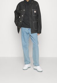Chaqueta de denim negra con herrajes dorados y costuras blancas, sobre un hoodie negro, combinada con jeans de color azul claro y zapatillas blancas.