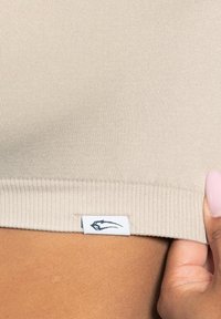 Brassière en tissu côtelé beige avec une étiquette blanche portant un logo noir au bas. Texture lisse et ajustement serré.