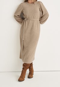 Beige stickad midi-klänning med långa, puffiga ärmar, midjeband och mjuk textur, i kombination med bruna knähöga stövlar i mocka.