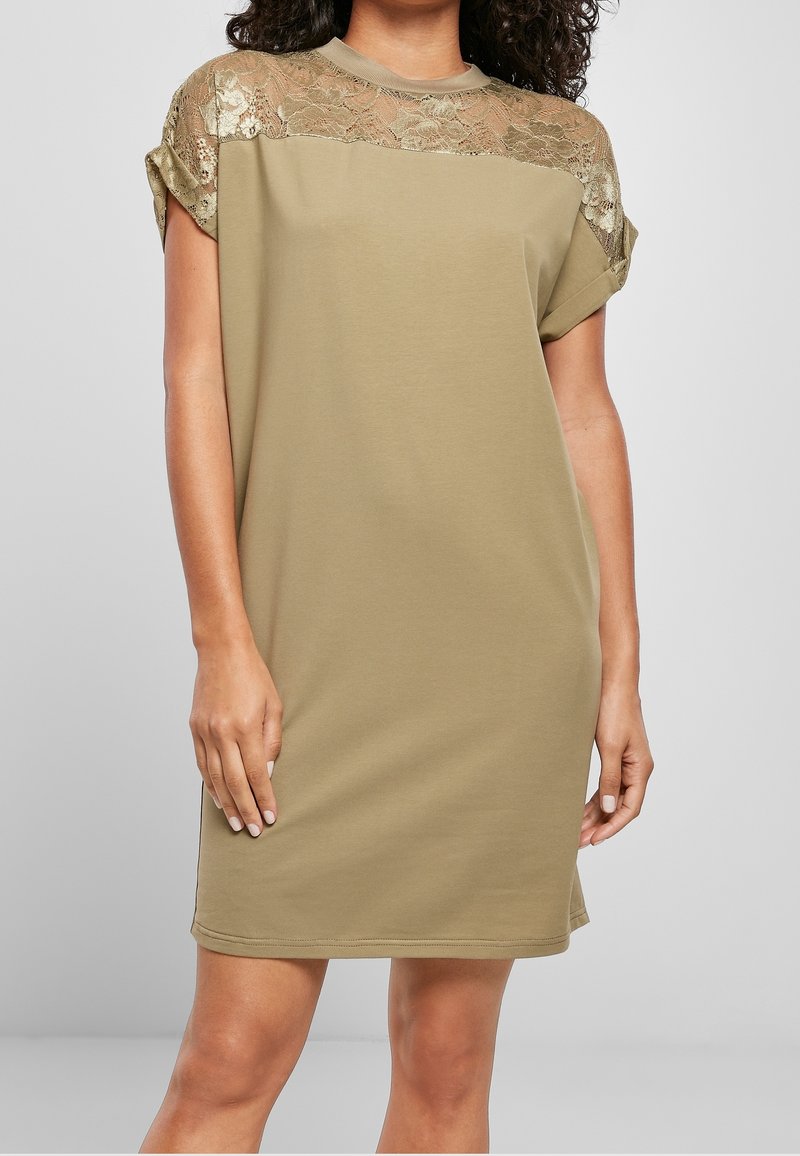 Femme portant une robe olive mi-longue avec un détail en dentelle sur le haut de la poitrine et des manches courtes retroussées.