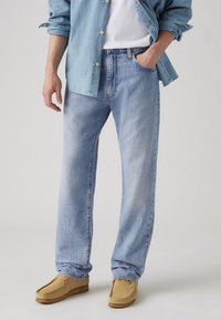 Pantalones vaqueros de mezclilla azul claro con un ajuste relajado, que cuentan con un diseño clásico de cinco bolsillos y un sutil desgaste. Combinados con zapatos mocasines en beige.