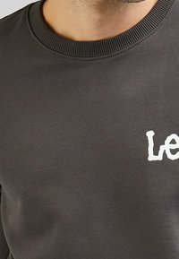 Grå sweatshirt med ribbad rundhals, med en vit tryckt logotyp på bröstet. Tyget verkar slätt och medelhäftigt.