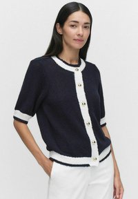 Stickad cardigan i marinblått med vita detaljer. Har korta ärmar, rund hals och guldtonade knappar framtill. Strukturert tyg.