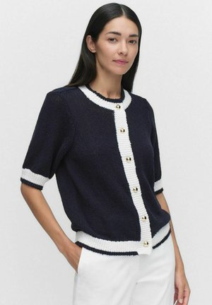 Stickad cardigan i marinblått med vita detaljer. Har korta ärmar, rund hals och guldtonade knappar framtill. Strukturert tyg.