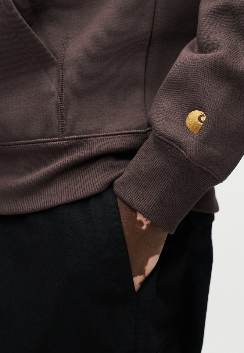 Brun sweatshirt med ribbede ærmegab, der har en guld logo detalje på ærmet. Sorte bukser med en synlig tekstureret lomme.