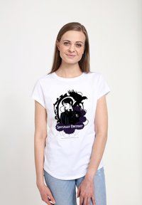 Žena v bílem tričku s černým a fialovým designem, na kterém je znázorněna žena, krkavec a květiny, s nápisem "Sociálně vzdálená", pózuje na bílém pozadí.