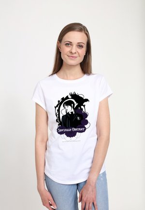 Kobieta w białym T-shircie z czarno-purpurowym wzorem przedstawiającym kobietę, kruka i kwiaty, z napisem "Społecznie Dystansująca", pozująca na tle białym.