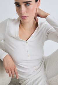 Kvinna med blå ögon och mörkt hår har på sig en vit knäppt långärmad topp och byxor, sitter avslappnat mot en ljus bakgrund.