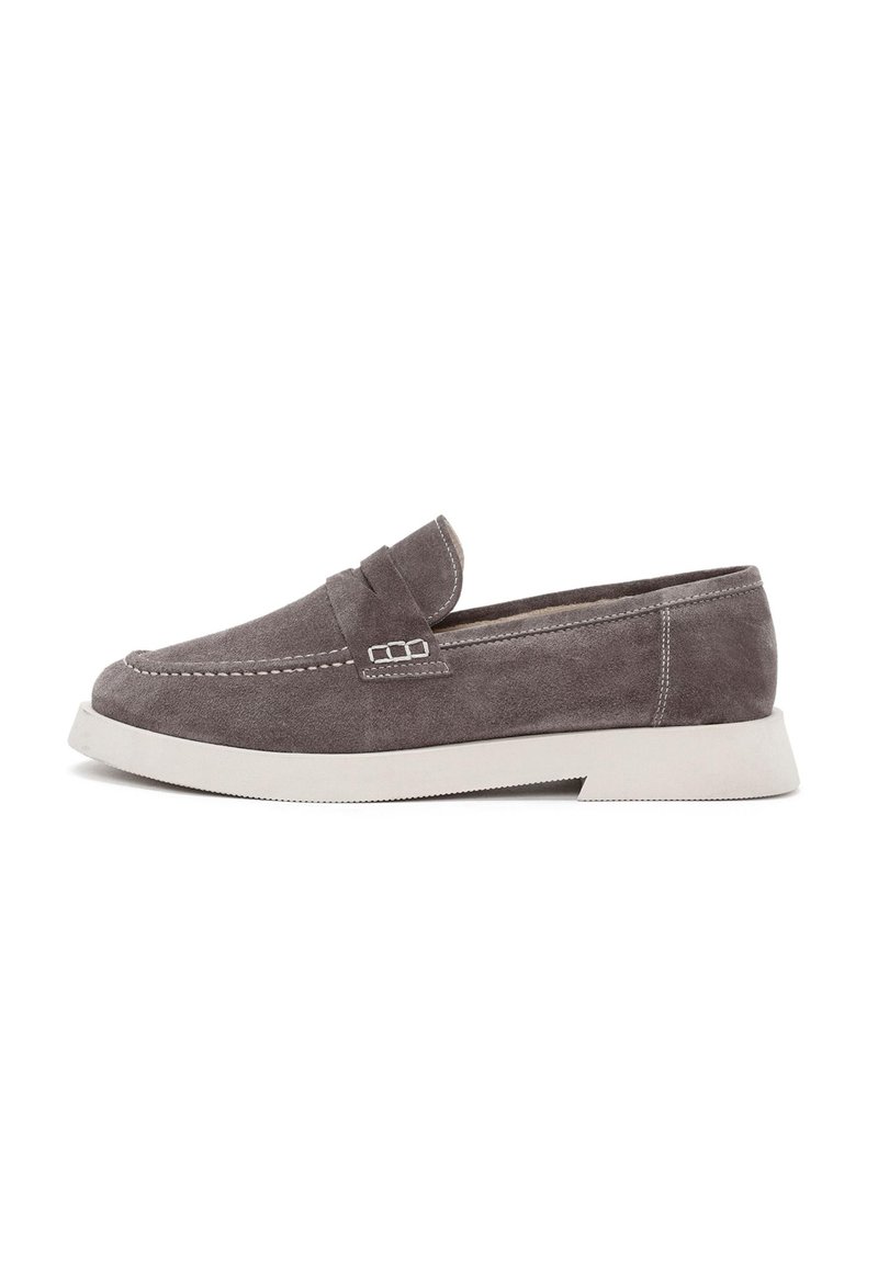 Mocasines de ante gris con una suela de goma blanca ligeramente elevada, que presentan un diseño cosido y un pequeño acento decorativo en la parte superior.