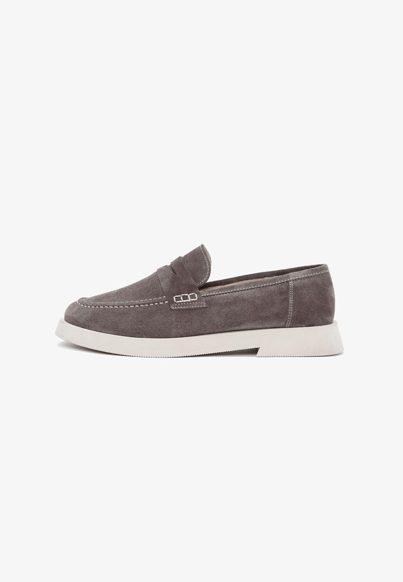 Mocasines de ante gris con una suela de goma blanca ligeramente elevada, que presentan un diseño cosido y un pequeño acento decorativo en la parte superior.