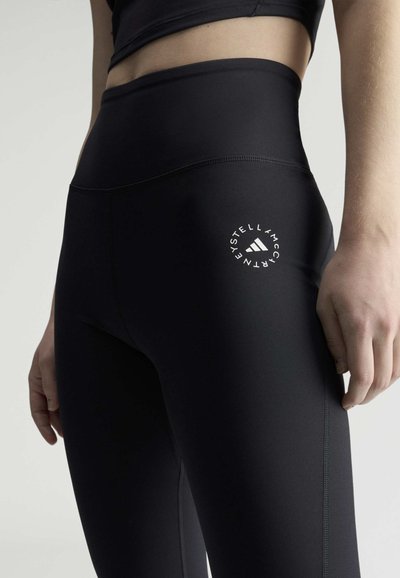 Nærbillede af en person iført sorte leggings med høj talje og en hvid, rund Adidas by Stella McCartney-logo på venstre lår.