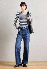 Gestreiftes Langarmshirt in Marineblau und Weiß, kombiniert mit hochtaillierten, weiten blauen Jeans. Das Model hält eine schwarze Handtasche und steht auf einem Holzfußboden.