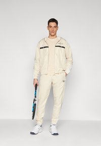 Vīriešu sporta apģērbs gaiši bēšā krāsā, kurā ietilpst kapuce ar melnām svītrām, pieguļošs krekls un pieguļoši bikses. Tur rokās tenisa raketi.