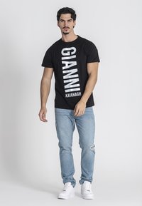 Čierne tričko s krátkym rukávom s veľkým bielym nápisom "GIANNI KAVANAGH", kombinované s svetlomodrými džínsami a bielymi teniskami. Pozadie je jednoduché.