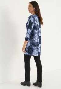 Blaue und marinefarbene Tunika mit Spritzerdesign, Dreiviertelärmel, kombiniert mit schwarzen Slim-Pants und Ankle-Boots. Glatte Stofftextur.