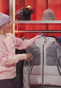 Person i rosa genser og lilla lue holder en grå og mørkegrå dunjakke på en kleshenger i en butikk med skiutstyr i bakgrunnen.