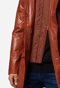 Bruine leren jas met een gladde textuur, voorzien van een gewatteerde binnenvoering, dubbele ritsen en geknoopte zijzakken.